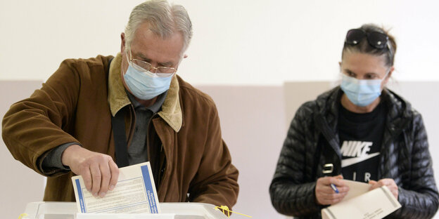 Mann mit Mund-Nase-Schutz steckt Stimmzettel in Urne, Frau wartet