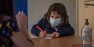 Ein Kind mit Mundschutzmaske und einem roten Stift in der Hand sitzt an seinem Schreibtisch