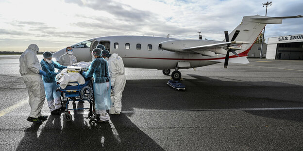 Menschen in Schutzanzügen schieben eine krankenliege vor einem kleinen Flugzeug