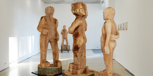 Große, grob gesägte Holzfiguren auf Sockeln stehen nebeneinander in einem Raum