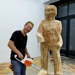 Ein Mann mit Säge steht neben einer Holzskulptur