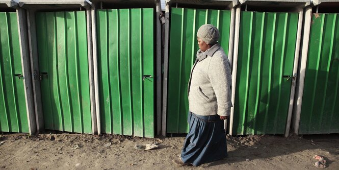 eine schwarze Frau geht an einer Reihe von öffentlichen Toiletten mit grüner Tür, vorbei