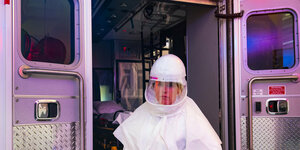 Eine Frau in Schutzkleidung steht vor den geöffneten Hintertüren eines Krankenwagen