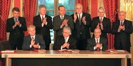 Franjo Tudjman und Slobodan Milošević und Alija Izetbegovic sitzen an einem Tisch, vor ihnen die Vertragsmappen. Hinter ihnen lauter wichtige Staatschefs, u.a. Bill Clinton und Helmut Kohl. Alle applaudieren