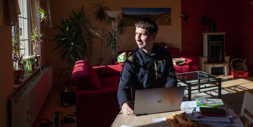 Gleichmann sitzt mit Laptop am Tisch im Wohnzimmer, hinter ihm sind rote Sofas zu sehen, die Sonne scheint seitlich in sein Gesicht