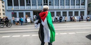 Ein Demonstrant mit einer Flagge Palästinas umhüllt steht während einer Demonstration in München auf der Straße.