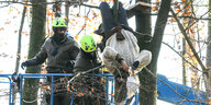 Eine Frau hängt kopfüber von einem Baumast. Zwei Männer mit gelbgrünen Schutzhelmen versuchen sie auf eine Hebebühne zu ziehen