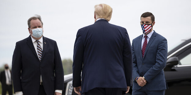 Trump von hinten, ihm gegenüber zwei Männer mit Mund/Nasenschutz
