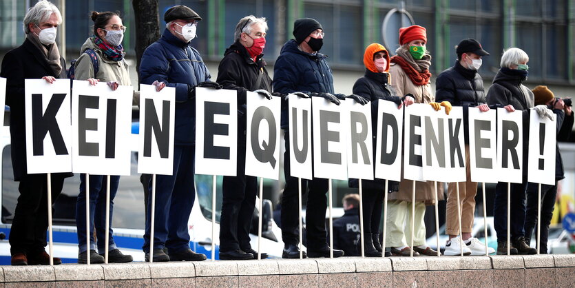 Mehrere Menschen mit Mund/Nasenschutz stehen auf einer Mauer, vor ihnen Schilder mit je einem Buchstaben, zusammen ergeben sie: Keine Querdenker!