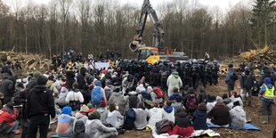 Mehrere Dutzend Menschen sitzen und stehen um einen Bagger herum, der von Polizei geschützt wird