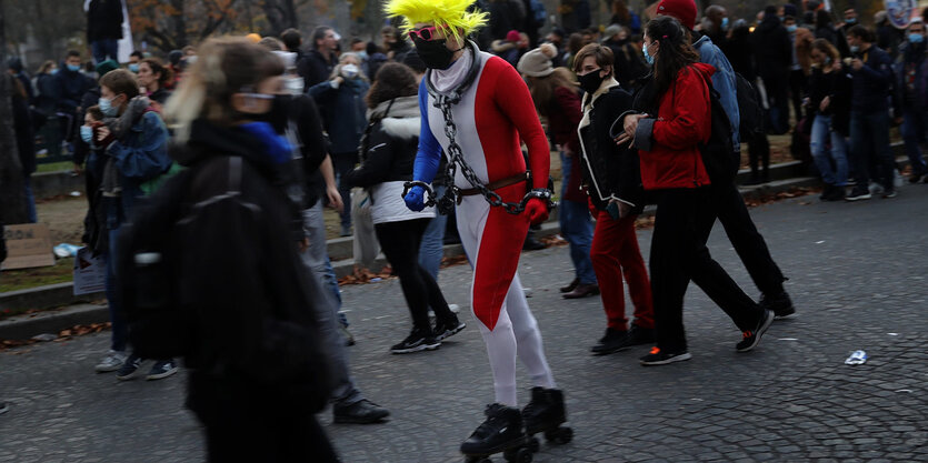 Ein Mann in einem Overall in den französischen Nationalfraben trägt Handfesseln un d eine Kette um den Hals. ERfährt auf Rollschuhen durch eine Menschenmenge