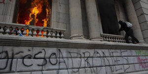 Aus einem Fenster des Parlamentsgebäudes lodern Flammen, eine vermummte Gestalt klettert an der Außenwand. Ein Graffito an der Wand lautet übersetzt "Diebe".