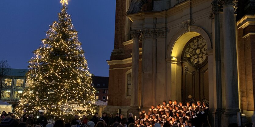 Ein Chor singt vor Kirche neben großem Tannnenbaum.