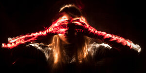 Fotografie der Künstlerin Susanne Emmermann. Eine Frau mit langen roten Handschuhen hält sich ihre Augen zu. Unter ihren Händen - an der Position der Augen - scheint eine Lichtquelle durch.
