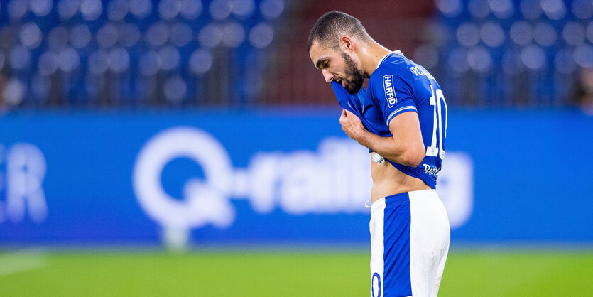 Schalker Fußballprofi wischt sich auf dem Rasen mit dem Trikot den Schweiß aus dem Gesicht