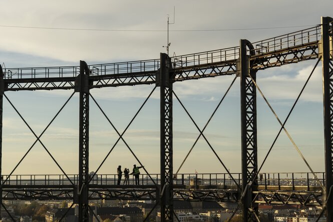 Personen stehen auf dem Industriedenkmal Schöneberg. Sie sind von weit aufgenommen, man erkennt nur ihre Silhouetten. Eine Person zeigt mit dem Arm in die Ferne, eine andere Person macht ein Foto.