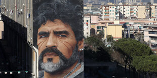 Ein Porträt von Maradona ist in riesigen Dimensionen an eine Hauswand gemalt, dahinter sieht man die Stadt Neapel