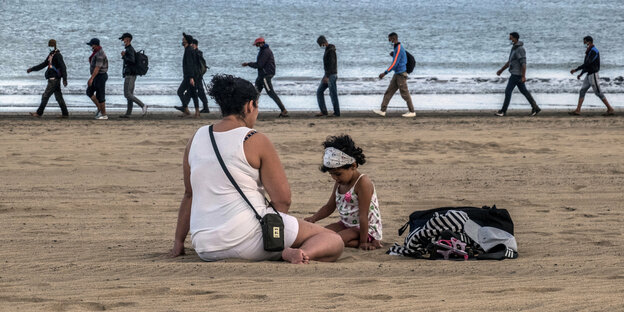 Migranten aus Marocco werden von der Polizei am Strand begleitet im Vordergrund eine Frau spielt mit Kind am Strand