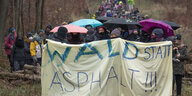 Aktivist*innen laufen mit Masken und Regenschirmen durch den Wald, im Vordergrund ein Banner mit der Aufschrift "Wald statt Asphalt"
