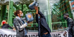 Menschen mit Masken halten Transparente und trinken aus Flaschen, die wie kleine Ölfässer mit Shel-Logo aussehen.