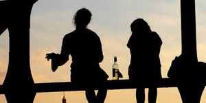 Silhouette von zwei Frauen, sie sitzen auf einer Brücke, zwischen ihnen steht eine Flasche WEin