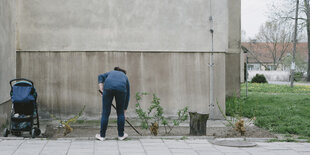 Ein Frau harkt vor einer grauen Betonhauswand ein Beet mit Sträuchern, neben ihr steht ein Kinderwagen