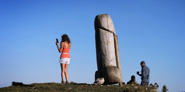 Der Holzpenis auf dem Hügel ist im Zentrum abgebildet, links davor steht eine Frau mit Handy und macht ein Selfie