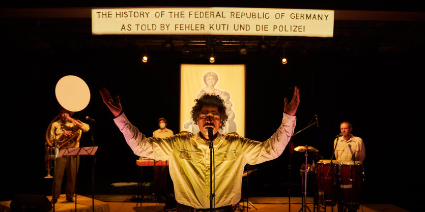 Fehler Kuti steht auf einer Bühne mit nach oben gereckten Armen, hinter ihm stehen Musiker und ein Madonnenbild hängt am Bühnenende. Darüber steht The History of the Federal Republic of Germany as told by Fehler Kuti und die Polizei