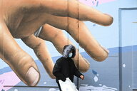 Ein Mann mit Kapuze und Schutzmaske läuft vor einem graffiti, das eine Große Hand zeigt
