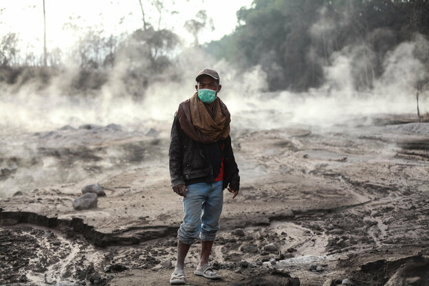 Ein Mann, der als vorbeugende Maßnahme gegen die Verbreitung des neuen Coronavirus einen Mund-Nasen-Schutz trägt, steht auf einer Fläche, die bedeckt ist mit vulkanischem Material des ausgebrochenen Vulkans Semeru und von der Rauch aufsteigt