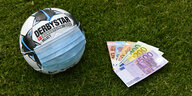 Ball mit übergestüpter Gesichtsmaske auf Rasen, daneben Geldscheine