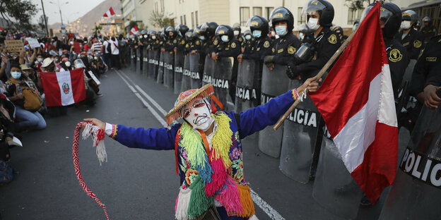 Ein Konstünierter Demonstrant mit peruanischer Flagge vor einer Reihe von Polizisten