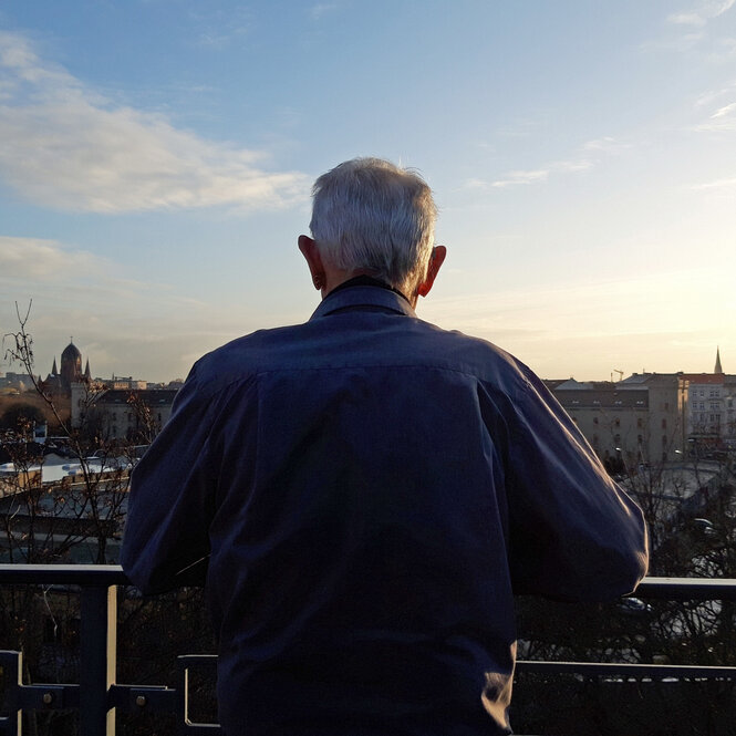 Peter G. ist nur von hinten zu sehen, er steht auf einem Balkon und blickt in den blauen Himmel