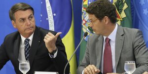 Die beiden Männer sitzen vor einem blauen Hintergrund. Bolsonaro deutet mit dem Daumen auf Salles.