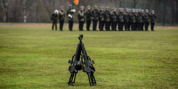 Sturmgewehre sind im Vordergrund auf einen Rasen aufgestellt. Unscharf im Hintergrund aufgereihte Soldaten der Bundeswehr