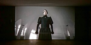 Eine Sequenz aus einem Film - ein Mann steht mystisch überhöht in einem schwarzen Kleid und hat eine Reitgerte in der Hand