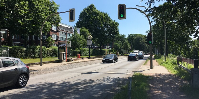 Eine Straße mit Autos, beidseits Bäume, über die Straße ragen Ampeln