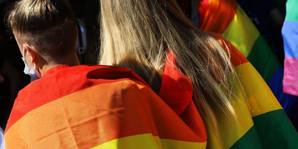 Zwei Frauen von hinten halten sich im Arm eingehüllt in einer Regenbogenflagge