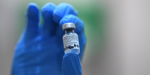 Eine Hand mit blauem Plastikhandschuh hält eine kleine Ampulle mit Impfstoff
