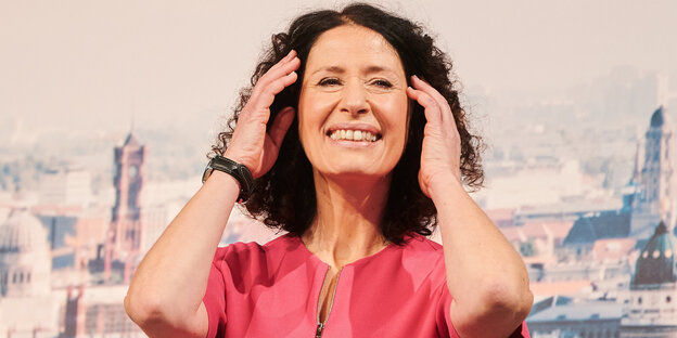 Das Bild zeigt die am Samstag zur Spitzenkandidatin gewählte Grünen-Politikerin Bettina Jarasch vor einer Berliner Stadtansicht. Sie ist eine Frau mittleren Alters mit lockigem Haar. Sie löchelt und greift sich in die Haare.