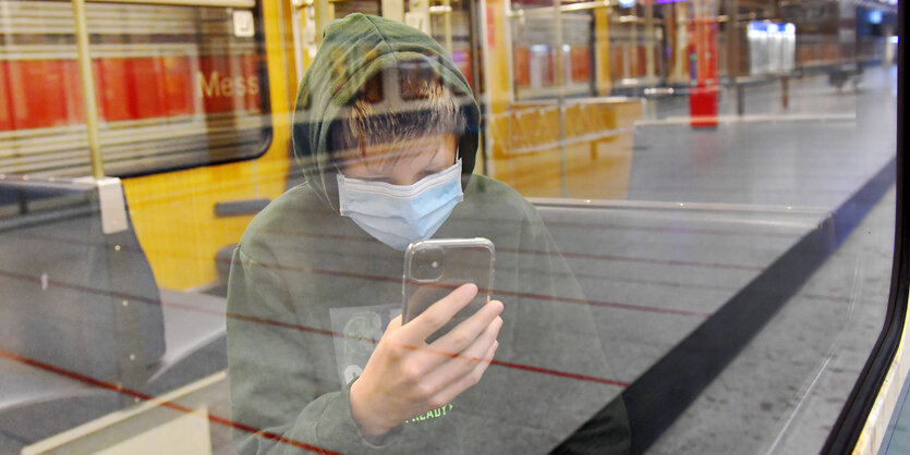 Ein Jugendlicher mit Mund-Nase-Maske sitzt in der Münchner U-Bahn und schaut auf sein Smartphone