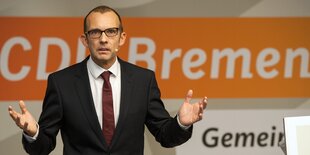 Thomas Röwekamp steht, in dunklem Anzug mit roter Krawatte auf weißem Hemd, vor einem CDU-Schriftzug (weiß auf orange) und winkelt die Arme an