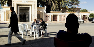Männer sitzen an einem Tisch auf einem Platz in Sidi Boutid. Im Hintergrund Palmen im Vordergrund eine Silhouette eines Mannes.