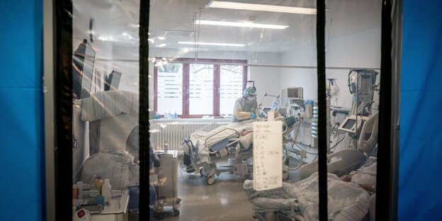 Ein Intensivpfleger ist hinter einer Schleuse bei seiner Arbeit in einer Schutzausrüstung mit Mund-Nasenbedeckung, Gesichtsschutz, Kittel und Haube auf der Intensivstation des Krankenhauses Bethel Berlin an einem Corona-Patienten zu sehen.