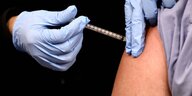 behandschuhte Hände verabreichen eine Impfspritze in einen Oberarm