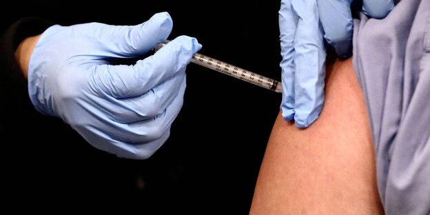 behandschuhte Hände verabreichen eine Impfspritze in einen Oberarm