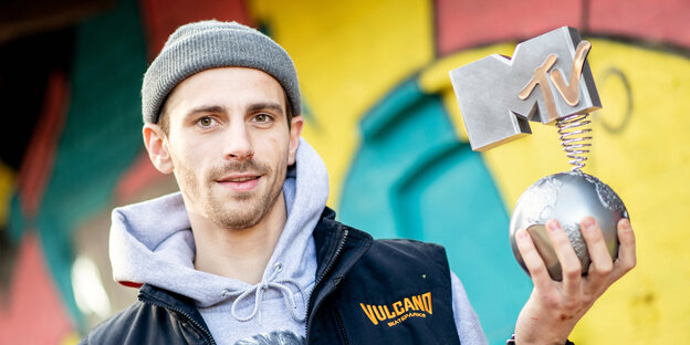 Mann mit Mütze hält eine MTV-Trophäe, im HIntergrund bunte Graffiti