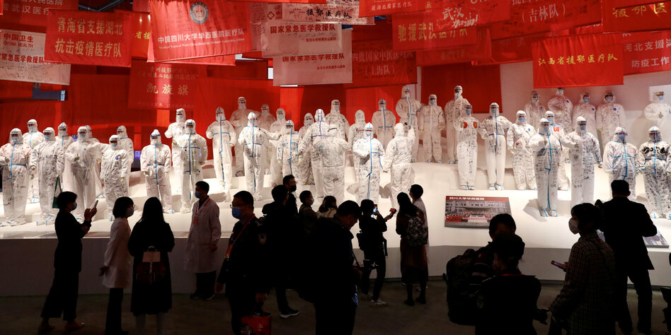 Auf einer Bühne mit rotem Hintergrund stehen mehrere Dutzend Menschen in komplett weißen Schutzanzügen, auch der Kopf und das Gesicht sind davon eingehüllt. Vor ihnen steht im Dunkeln Publikum