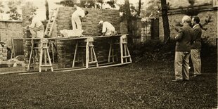 Zwei elegant gekleidete Männer stehen in einem Garten und beobchten die Maurer die eine Wand hochziehen