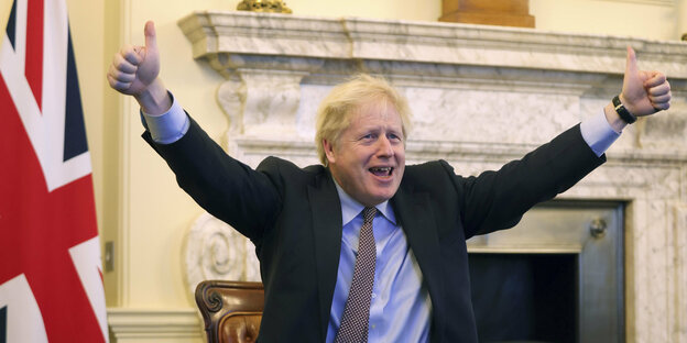 Boris Johnson, ein Mann mit wirren, blonden Haaren jubelt und reckt beide Daumen in die Luft. Im Hintergrund ein Kamin und die Flagge Großbritanniens